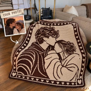 May include: A rectangular crochet blanket featuring a dark brown and cream-colored graphic design of two people embracing. The border is decorated with a repeating pattern of small hearts. A small inset photo shows the original reference image with text.