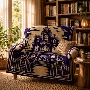 May include: A woven throw blanket featuring a dark purple and cream graphic design of a Victorian-style mansion with a gated entrance, bare trees, and bats against a full moon. The blanket is draped over an armchair with a small beige pillow.