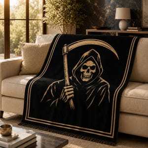 Könnte beinhalten: Eine schwarze Wohndecke mit einer goldfarbenen Grafik eines Skeletts mit Kapuze, das eine Sense hält. Das Motiv ist von einem doppelten Linienrahmen umgeben und weist am unteren Rand des rechteckigen Textilstücks Fransen auf.