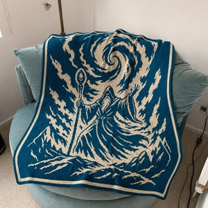 May include: A rectangular knit blanket featuring a two-tone design in deep teal and cream. The central graphic depicts a robed figure holding a tall staff, standing amidst a mountainous landscape with a swirling vortex pattern in the sky above.