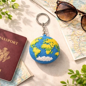 May include: A blue and yellow crocheted globe keychain with a silver keyring. The keychain is next to a passport, sunglasses, and a map. The globe features continents in yellow and green, with a white cloud at the bottom.