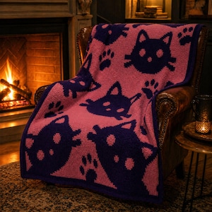 May include: Pink and navy blue cat-themed throw blanket. The blanket features a repeating pattern of cat faces and paw prints. The blanket is draped over a leather armchair in front of a fireplace.