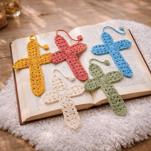 May include: Five crochet cross bookmarks in yellow, coral, cream, green, and blue rest on an open book. Each cross has a loop at the top. The book is open on a white, fluffy surface.