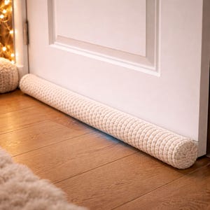 Puede incluir: Un tope de puerta cilíndrico de color crema, hecho de tela de punto. El tope está colocado en la base de una puerta blanca, apoyado en un suelo de madera. La textura de punto es visible y el tope mide aproximadamente 91 cm de largo.