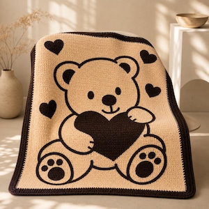May include: A tan and brown crocheted blanket featuring a large teddy bear holding a heart. The blanket has a dark brown border and several heart accents. The teddy bear design is outlined in dark brown.