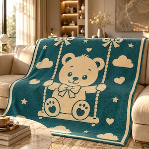 May include: A teal and cream-colored knit blanket featuring a central graphic of a teddy bear sitting on a swing. The design includes decorative elements such as small stars, hearts, and clouds surrounding the bear, with a bow tie on the bear.