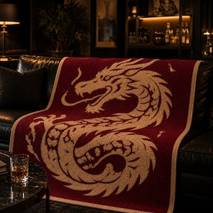Op de afbeelding: Een rode plaid met een gouden drakenontwerp, gedrapeerd over een zwarte leren bank. De draak is gedetailleerd, met een lang lichaam en open mond. De deken heeft een getextureerd uiterlijk en de achtergrond is zwak verlicht.