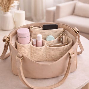 May include: A beige leather handbag with a woven organizer insert. The organizer holds various items, including a pink jar, small bottles, a compact, keys, and a smartphone. The bag has a shoulder strap and gold-colored hardware.