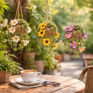 May include: Three hanging crochet flower baskets with white, yellow, and purple flowers. The baskets are suspended by twine and are filled with green foliage. A white coffee cup and sunglasses sit on a wooden table in the foreground.
