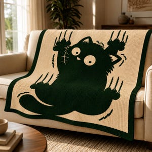 Peut inclure: Un plaid tricoté rectangulaire présentant un grand motif vert foncé représentant un chat aux yeux écarquillés avec les griffes sorties, sur un fond beige clair. La couverture possède une bordure vert foncé unie encadrant l'illustration.