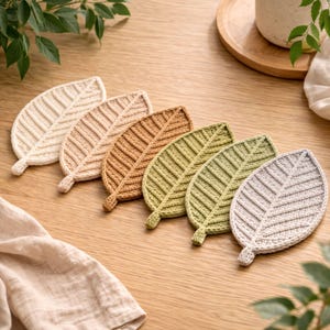 Puede incluir: Un conjunto de seis posavasos con forma de hoja en varios colores neutros, incluyendo crema, beige y tonos de marrón y verde. Los posavasos están hechos de un material tejido con textura y están colocados sobre una superficie de madera.