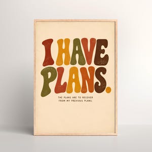 May include: A framed wall art print featuring the text I HAVE PLANS. in large, wavy, multi-coloured letters of red, yellow, green, and brown. Below this, smaller black text reads THE PLANS ARE TO RECOVER FROM MY PREVIOUS PLANS. The print is set in a light wood frame.