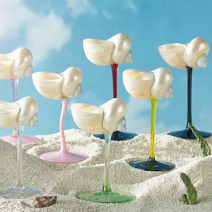 Peut inclure: Sept verres à vin originaux avec des coupes en forme de coquillage et des pieds colorés. Les pieds sont transparents, roses, rouges, jaunes et noirs. Les verres sont disposés sur du sable, avec un ciel bleu en arrière-plan.