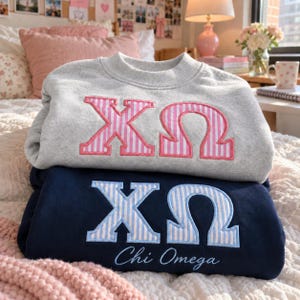 Op de afbeelding: Een lichtgrijze sweater met ronde hals en roze-wit gestreepte Griekse letters Chi Omega, liggend op een marineblauwe sweater met ronde hals met lichtblauw-wit gestreepte Griekse letters en de tekst Chi Omega in script.