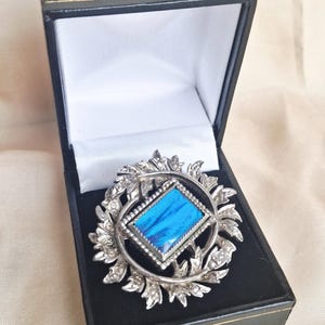 May include: A silver brooch featuring a central blue rectangular stone, encircled by a decorative frame. The brooch is set within a wreath of leaves and crystals, displayed in a black jewellery box with a white lining.