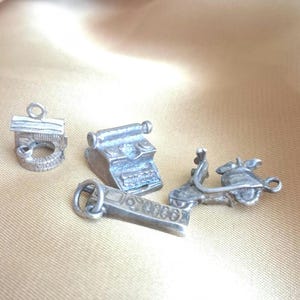 May include: A collection of small, silver-toned charms. The charms include a well, a typewriter, a scooter, and a rectangular charm with engraved details. The charms are displayed on a gold-colored fabric.