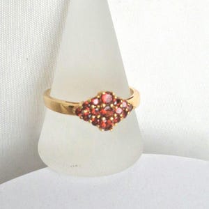 May include: A gold ring featuring a diamond-shaped cluster of small, round, red gemstones. The ring is presented on a clear, cone-shaped stand against a white backdrop. The ring's band is plain, polished gold.
