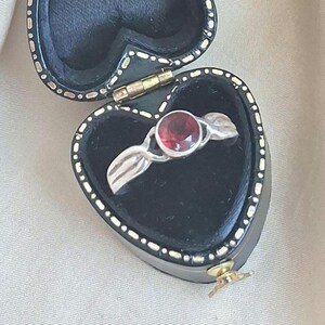 May include: A silver ring featuring a round, red gemstone, with a leaf-shaped design. The ring is presented in a black, heart-shaped jewellery box with a gold clasp and white dotted trim.