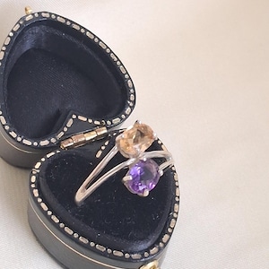 May include: A silver ring featuring two oval-cut gemstones, one light orange and one purple, set in a bypass style. The ring is displayed inside a small, black, heart-shaped jewelry box with a black velvet interior and gold-tone metal trim.