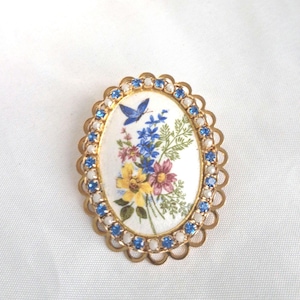 May include: Oval-shaped brooch with a gold-tone scalloped edge, adorned with blue gemstones. The center features a floral design with a blue butterfly. The flowers are in shades of yellow, blue, and purple.