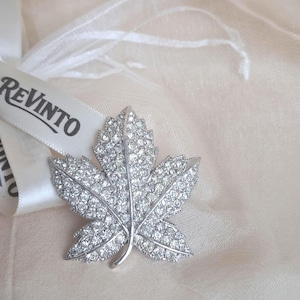 May include: A silver-coloured leaf-shaped brooch, adorned with many clear crystals. The brooch is set against a soft, cream-coloured fabric. The word "REVINT0" is printed on a white ribbon.