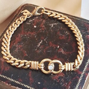May include: A gold bracelet with a curb chain design. The bracelet has a central feature of two interlocking circles with a small, clear gemstone. The clasp is a lobster claw style.