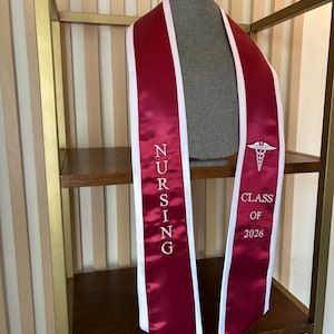 May include: A burgundy satin graduation sash with white trim, draped over a mannequin. The sash is embroidered with the word "NURSING" vertically and "CLASS OF 2026" along with a medical symbol. The sash is displayed on a wooden shelf.