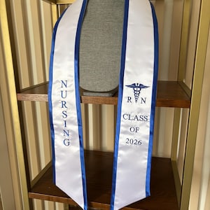 May include: White graduation sash with blue trim and text. The sash reads "NURSING" vertically and "RN CLASS OF 2026" with a caduceus symbol. The sash is displayed on a mannequin head.