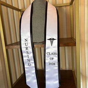 May include: White satin graduation sash with black edging. The sash displays the word "NURSING" in vertical black letters on one side and the medical caduceus symbol with "CLASS OF 2026" on the other side.