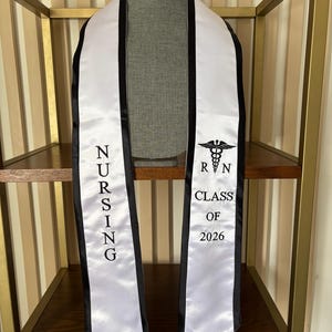 May include: White graduation sash with black trim and the text "NURSING" on one side and "RN CLASS OF 2026" with a caduceus symbol on the other. The sash is displayed on a mannequin.