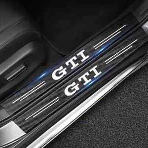 Könnte beinhalten: Schwarze Carbonfaser-Autotürschwellerschützer mit dem Wort "GTI" in weißen Blockbuchstaben. Jeder Schutz hat zwei parallele weiße Linien. Sie sind in einem Autoinnenraum installiert.