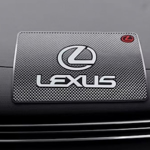 以下が含まれることがあります： 白と赤のレクサスロゴが入った長方形の車のダッシュボードマット。マットはテクスチャード加工された幾何学模様で、レクサスの名前が白いブロック文字で表示されています。