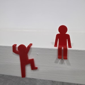 Peut inclure: Deux sculptures stylisées de personnages humains rouges. Une figurine grimpe, l'autre est debout. Les figurines sont placées sur une surface blanche, avec un fond blanc uni.