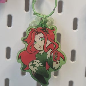 May include: A clear acrylic keychain featuring a cartoon illustration of a character with long red hair and green eyes, wearing a green leafy outfit. The character is adorned with small green sprigs in their hair and hand. It hangs from a light green ring.