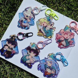 Könnte beinhalten: Eine Sammlung farbenfroher Acryl-Schlüsselanhänger mit Anime-Charakteren. Jeder Schlüsselanhänger hat ein einzigartiges Design mit leuchtenden Farben und ist an einem Metallverschluss befestigt. Die Schlüsselanhänger liegen auf einer weißen Oberfläche auf grünem Gras.