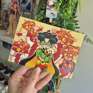 Puede incluir: Una impresión artística rectangular con una ilustración de dibujos animados de un personaje de superhéroe en rojo, verde y amarillo. La impresión tiene un fondo amarillo con múltiples figuras en poses de acción. La impresión está sostenida por una mano.
