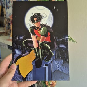 May include: A comic book illustration of a superhero, perched on a rooftop under a full moon. The character wears a red and black costume with a yellow cape. The background depicts a cityscape at night.