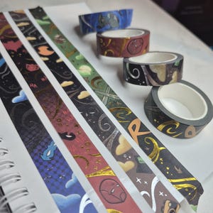 Könnte beinhalten: Dekorative Washi-Tape-Rollen und -Streifen mit verschiedenen Designs. Das Tape zeigt Illustrationen von Charakteren, Wolken und Mustern in Farben wie Blau, Rot, Grün und Gold. Die Rollen werden neben Streifen der gleichen Designs präsentiert.