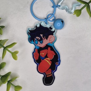 May include: A blue keyring featuring a cartoon character in a Superman-inspired outfit. The character has black hair and sunglasses. The keyring includes a blue ring, a star-shaped charm, and a small blue bell. The character is giving a thumbs-up.