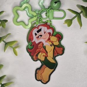 Pode incluir: Um chaveiro verde com um personagem de desenho animado com cabelo ruivo, uma roupa amarela e verde e uma mecha branca no cabelo. O chaveiro tem um pingente verde em forma de flor e um sino verde.