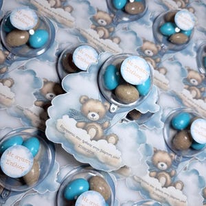 Könnte beinhalten: Transparente Kunststoffbehälter gefüllt mit blauen und beigen Bonbons, befestigt an wolkenförmigen Karten mit einer braunen Teddybär-Illustration und dem griechischen Text 'Ευχαριστώ που ήρθατε στην βάπτιση μου' sowie dem Datum 27/09/2026.