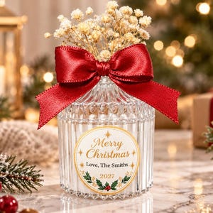 Peut inclure: Un bocal en verre transparent cannelé rempli de fleurs blanches séchées, orné d'un nœud en ruban de satin rouge foncé. Une étiquette circulaire porte le texte doré Merry Christmas, Love, The Smiths, 2027, avec des motifs de houx et d'étoiles.