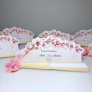 Può includere: Un ventaglio pieghevole con manico in legno chiaro e foglio in carta bianca con un'illustrazione floreale rosa, strass trasparenti e il testo stampato Forever & Always, Jack, Eleanor e la data 12.12.26, arricchito da un fiore in tessuto rosa.