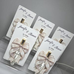 May include: Several wedding favor cards with perfume bottles. Each card features a cream-colored bow with gold glitter accents and a gold-capped perfume bottle. The cards are labeled "Jennifer & Thomas" and "Our Wedding 06.21.2026".