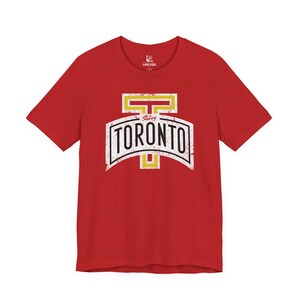 Può includere: T-shirt rosso con grafica bianca e nera invecchiata con la scritta "TORONTO" in stile vintage. La grafica include anche dettagli gialli e la parola "Savvy".