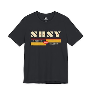 Peut inclure: T-shirt noir avec un motif graphique représentant le mot "SUSY" dans une police vintage. En dessous, les mots "GALLETA" et "RELLENA", avec un motif rectangulaire rouge et jaune entre les deux. Le t-shirt a un col rond classique.