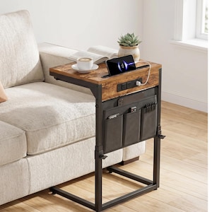 May include: A brown and black side table with a wooden top and metal frame. The table has a built-in charging station, a storage pocket, and is positioned next to a beige sofa. A phone, coffee cup, and book are on the table.