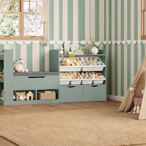 May include: A sage green wooden toy storage unit featuring a bench seat with a gray cushion, open shelving, and four white plastic bins filled with various toys. The unit includes two lower drawers and a top shelf holding books and decorative items.