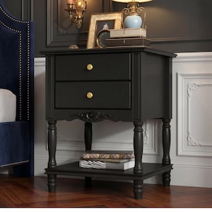 May include: A black wooden nightstand featuring two drawers with round gold-toned knobs, a carved floral detail on the front apron, four turned legs, and an open bottom shelf holding two stacked books. The piece has a smooth matte finish.