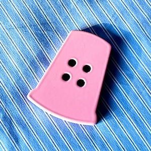 Peut inclure: Un bouton rose en céramique avec quatre trous noirs, posé sur un tissu à rayures bleues et blanches. Le bouton est de forme trapézoïdale avec des bords arrondis.